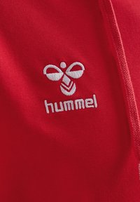 Felpa con cappuccio rossa in tessuto, caratterizzata da un logo bianco ricamato con un design stilizzato sopra la parola "hummel", che enfatizza la texture e i colori vivaci.