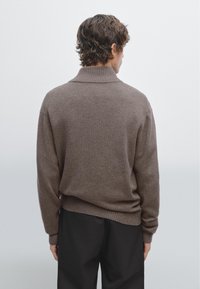 Brauner Strickpullover mit hohem Kragen und gerippten Bündchen. Lockerer Sitz, weiche Textur und einfaches Muster. Über dunkler Hose getragen.