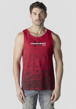 Hombre de pie mirando hacia adelante, vistiendo una camiseta sin mangas roja con patrón negro desvaído y texto "KOROSHI BRAND" y pantalones cargo gris claro.