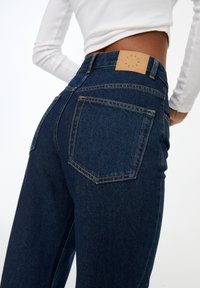 Jeans de mezclilla en azul oscuro con un talle alto. Presentan bolsillos traseros visibles, costuras en contraste y un parche de cuero marrón en la cinturilla.