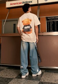 Camiseta de gran tamaño color beige con un gráfico de un oso en la parte posterior, combinada con jeans de pierna ancha azul deslavado y zapatillas blancas gruesas.