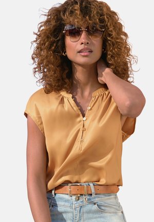 Woman with curly hair wearing sunglasses, a golden satin blouse, light blue jeans, and a brown belt with a gold buckle, hand touching neck.