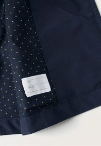 Marinblått tyg med ett prickigt mönster i guld. Inkluderar en vit etikett för namn och telefonuppgifter i hörnet.