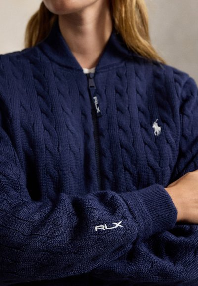 Personne portant un pull zippé en maille torsadée bleu marine avec le logo RLX sur la manche et un emblème blanc de joueur de polo sur la poitrine, bras croisés.