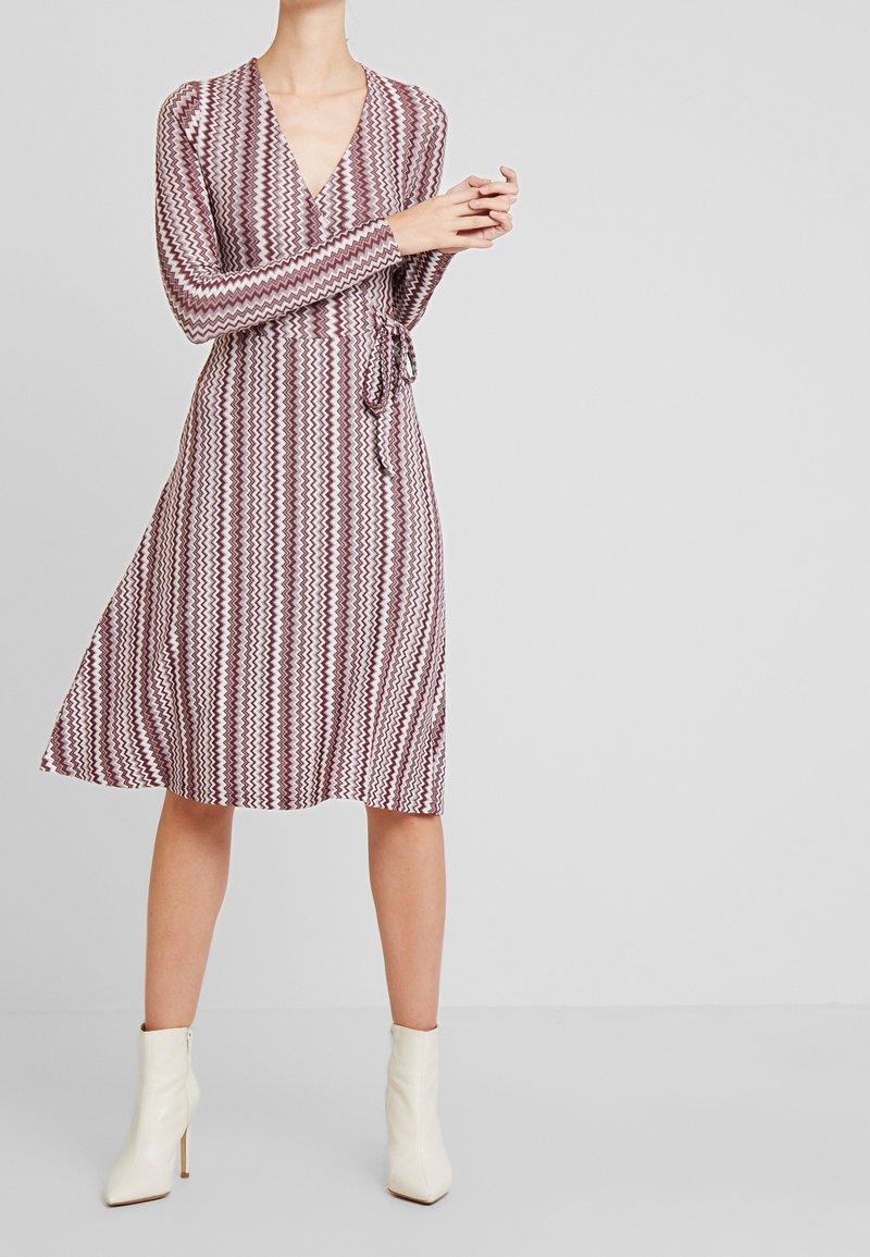 Femme portant une robe portefeuille mi-longue à manches longues avec des motifs en zigzag bordeaux et blanc, associée à des bottines blanches.