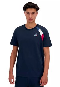 T-shirt a maniche corte di colore blu navy con colletto a girocollo, accenti triangolari rosso, bianco e blu sulla spalla e un logo sul petto.