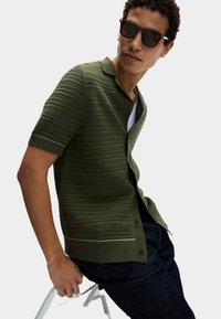 Camisa corta de manga oliva con rayas texturizadas, cierre de botones y cuello. Llevada sobre una camiseta blanca con jeans de mezclilla azul oscuro.