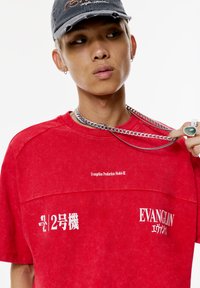 Tricou roșu oversized cu un finisaj texturat, având text imprimat alb "Evanglion Production Model-02" și "2号機" pe față.