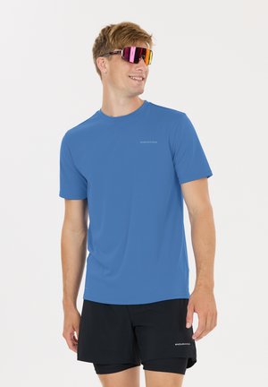 Homme portant une chemise à manches courtes bleue et un short noir, regardant sur le côté, avec des lunettes de sport réfléchissantes.