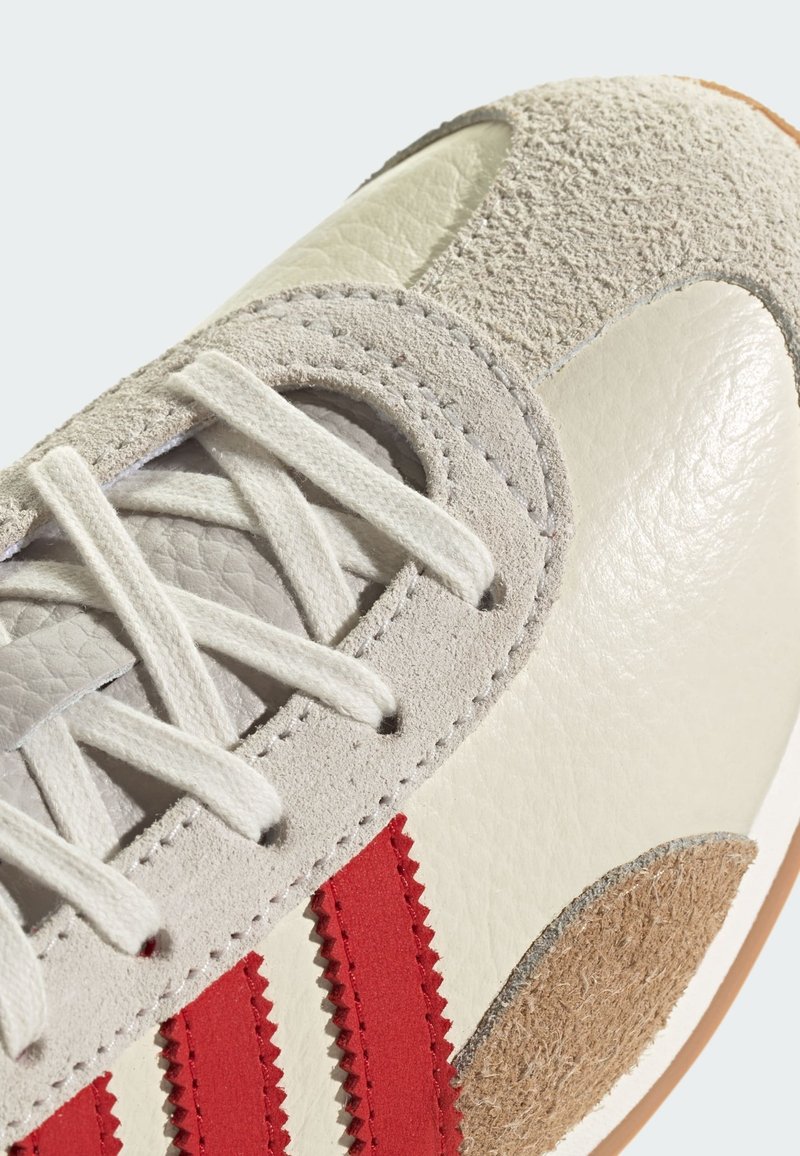 Zapatillas de cuero y gamuza en color crema, con acentos de rayas rojas y secciones texturizadas. Diseño con cordones y detalles cosidos.