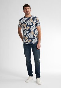 Lyhythihainen tummansininen paita, jossa on vaaleansininen ja kerma koristeellinen kuvio, yhdistettynä tummiin farkkuihin ja valkoisiin lenkkareihin.