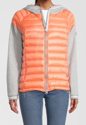 Kobieta ubrana w kurtkę na zamek błyskawiczny z błyszczącym, pomarańczowym, pikowanym przodem oraz jasnoszarymi rękawami i kapturem, w połączeniu z niebieskimi dżinsami.