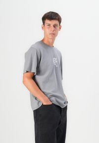 Grå kortærmet T-shirt i blødt stof med rund halsudskæring. Har et subtilt drageprint på brystet og en afslappet pasform, parret med mørke bukser.