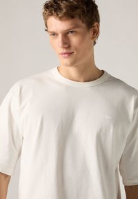 Λευκό μπλουζάκι από απαλό βαμβάκι, με στρογγυλή λαιμόκοψη και κοντά μανίκια. Σημαντική λεπτομέρεια: μικρό λογότυπο στο στήθος. Λεία υφή.