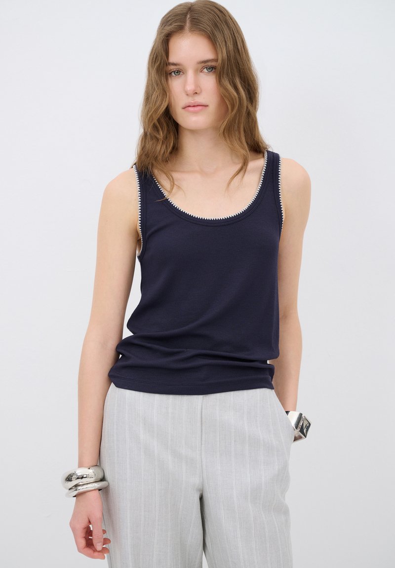 Canotta senza maniche blu navy con texture a coste e cuciture bianche a contrasto attorno al colletto, abbinata a pantaloni a righe grigio chiaro.