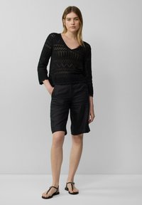 Schwarzes Strickoberteil mit V-Ausschnitt und Dreiviertelärmeln, kombiniert mit schwarzen Leinen-Shorts. Die Sandalen haben dünne Riemen. Einfaches, vielseitiges Outfit.