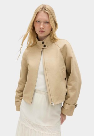 Lyshåret kvinde iført en beige lynlåstrøje med knappet krave og manchetter over en hvid kjole med blonder, foran en ensfarvet baggrund.