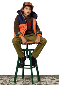 Un garçon portant un gilet orange et bleu marine, un pull marron, un pantalon vert, des baskets noires et une casquette, assis sur un tabouret vert sur un tapis à motifs.