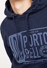 Osoba v námořnickém modrém hoodies s velkým logem "Imported Bell" a šňůrkami visícími vpředu.