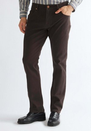 Mann trägt dunkelbraune gerade geschnittene Hose mit Knopfleiste, schwarze Lederloafers und ein gestreiftes Langarmshirt.