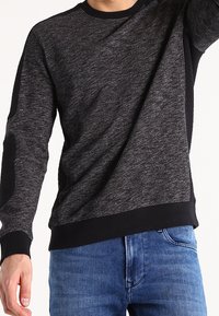 Sweatshirt gris foncé chiné avec des accents noirs. Doté d'un col rond et d'un tissu texturé sur les manches. Porté avec un jean bleu.
