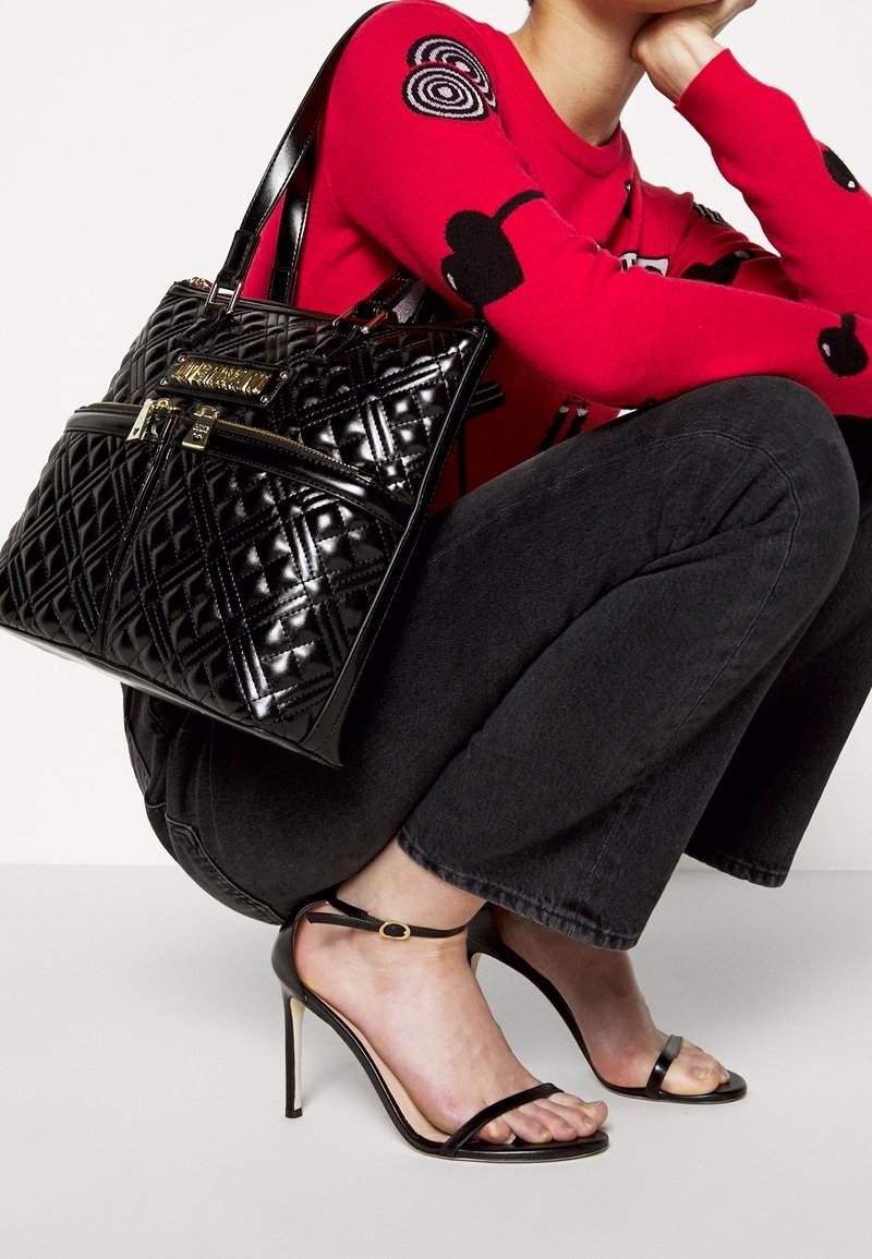 Personne portant des talons aiguilles noirs à brides, un jean noir, un pull rouge avec des motifs noirs, tenant un sac à main matelassé noir avec des détails de fermeture éclair dorés.