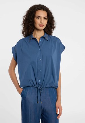 Frau mit lockigem Haar, die ein blaues kurzärmeliges Hemd mit Schmuckkragen und passende blaue Hosen mit Kordelzug trägt, Hände in den Taschen.