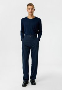 Marineblauwe langetermijnshirt gecombineerd met hoge marineblauwe broeken en zwarte schoenen. De outfit heeft een getailleerd ontwerp en een strakke riem.