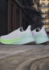Paire de chaussures de course blanches et vert clair placées dans une rue de la ville avec des bâtiments en arrière-plan.