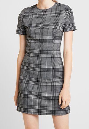 Femme portant une robe mini ajustée à manches courtes à motif plaid noir et blanc, se tenant devant un fond uni.