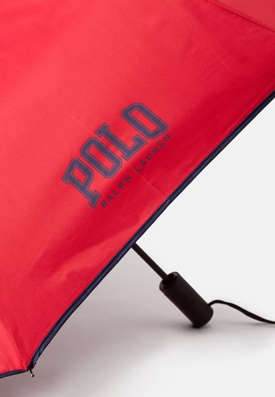Guarda-chuva vermelho com um contorno azul, apresentando um logótipo proeminente a dizer "POLO RALPH LAUREN." A textura do tecido parece suave e resistente à água.