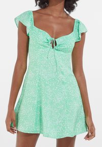 Green dress with white speckled pattern, featuring a square neckline, ruffled sleeves, and a tie detail at the bust. Flared silhouette.