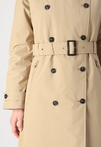 Manteau beige en trench avec un design double boutonnage, boutons noirs, ceinture avec boucle carrée et une poche latérale zippée. Texture lisse.