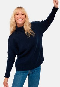 Dunkelblaues Strickpullover mit Stehkragen, entspannter Passform, überschnittenen Schultern und geripptem Saum, kombiniert mit hellblauen Jeans.