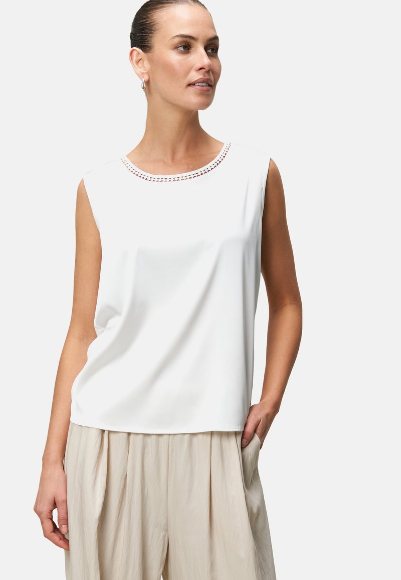 White sleeveless blouse with a round neck featuring decorative stitching. Made of smooth fabric, paired with light beige pleated trousers.
