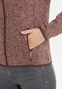 Fleecejacka i vinröd med ett strukturerat mönster, som har en ficka med dragkedja och åtsittande ärmar. Slät tyg och kontrasterande detaljer.
