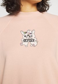 Rosa sweatshirt med en grafik av två tecknade kaniner som håller ett "LEVI'S"-skylte, med blommiga prydnader på sina huvuden. Mjuk textur.