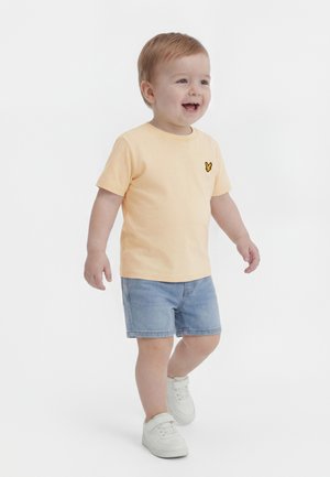 Tout-petit marchant, portant un t-shirt pêche avec un logo jaune, un short en denim et des baskets blanches, souriant et regardant devant lui.