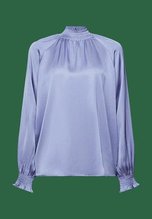 Blouse en satin lavande avec col montant smocké, manches longues bouffantes et poignets smockés sur fond vert.