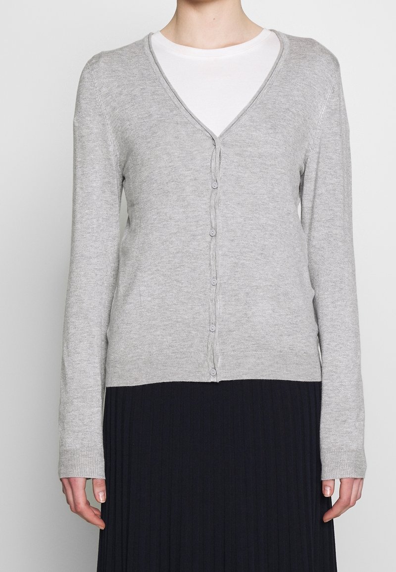 Personne portant un cardigan gris clair à col en V boutonné par-dessus un t-shirt blanc et une jupe plissée noire.