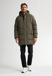 Manteau rembourré vert olive avec capuche, doté d'une fermeture éclair à l'avant, de deux poches latérales et d'un design mi-long. Porté avec un jean noir et des baskets.