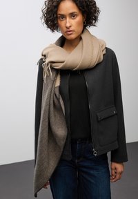 Sort cropped jakke med lynlås foran og lomme, parret med et beige uld tørklæde. Modellen bærer en sort top og blå denim jeans.