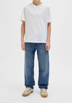 T-shirt en coton blanc, coupe ample. Jean en denim bleu foncé, coupe droite. Baskets beiges avec des accents bruns. Tenue simple et décontractée sur un fond uni.