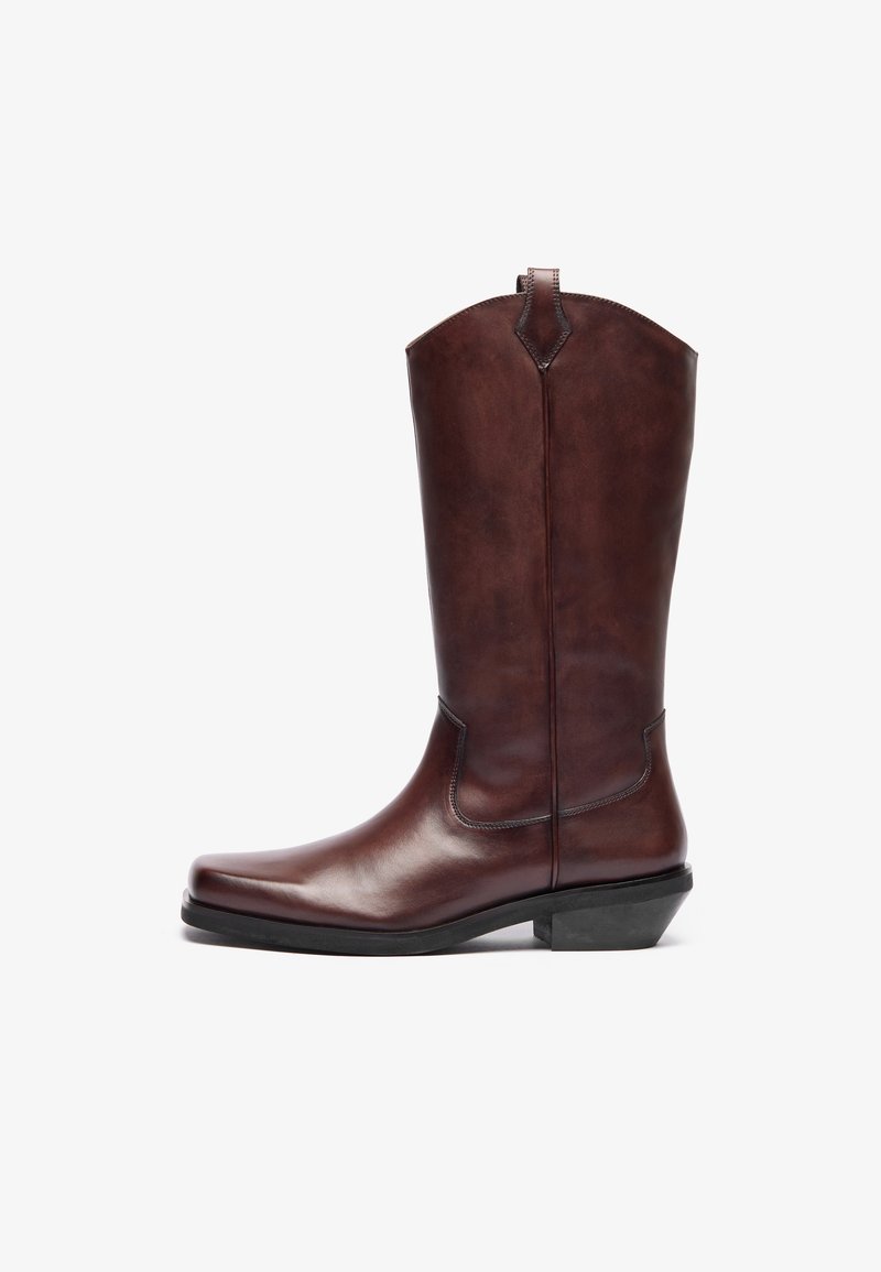 Bota de cowboy de pele castanha com bico pontudo, cano alto, textura suave e um ligeiro salto. Apresenta detalhes de costura discretos.