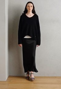 Černý oversized pletený svetr s dlouhými rukávy, vrstvený přes bílé triko, doplněný splývavou černou saténovou maxi sukni a modrými sandály na podpatku.