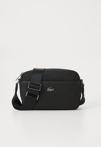 Czarna torebka typu crossbody wykonana z teksturowanej tkaniny, z zamkiem błyskawicznym, regulowanym paskiem i srebrnym logo Lacoste z przodu.