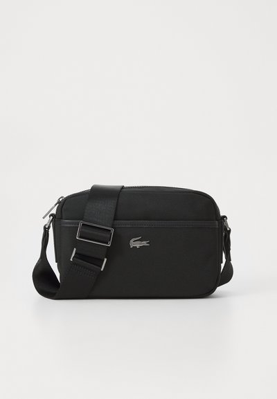 Sac bandoulière noir en tissu texturé, avec une fermeture zippée, une bandoulière réglable et un logo Lacoste en métal argenté sur le devant.