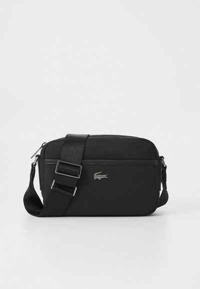 Sac bandoulière noir en tissu texturé, avec une fermeture zippée, une bandoulière réglable et un logo Lacoste en métal argenté sur le devant.