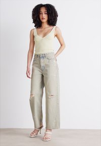 Light yellow ribbed tank top paired with distressed, oversized light blue denim jeans featuring rips. Model wears nude high-heeled sandals.