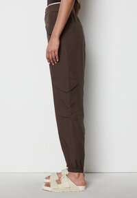 Braune Cargo-Hosen mit einem elastischen Bund, seitlichen Taschen und Bündchen. Kombiniert mit beigen Sandalen mit Schnallenakzenten. Flache Textur.